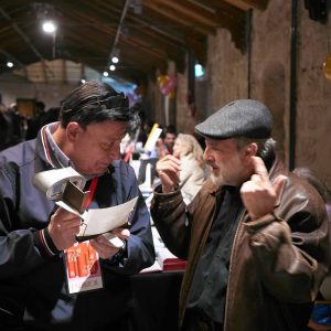 Close-up of two men talking at an art exhibition, one holding a brochure and the other gesturing. Art and stereography featured; capturing candid moments of interaction.
