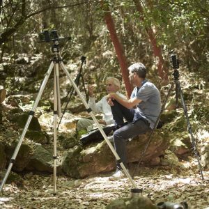 Close-up of two men sitting on rocks in a dense forest with professional camera equipment set up for stereography.